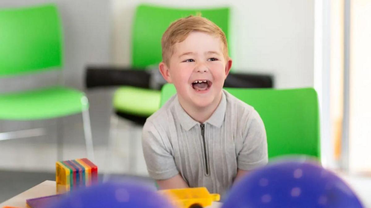 A young boy laughs at someone off camera, sitting down