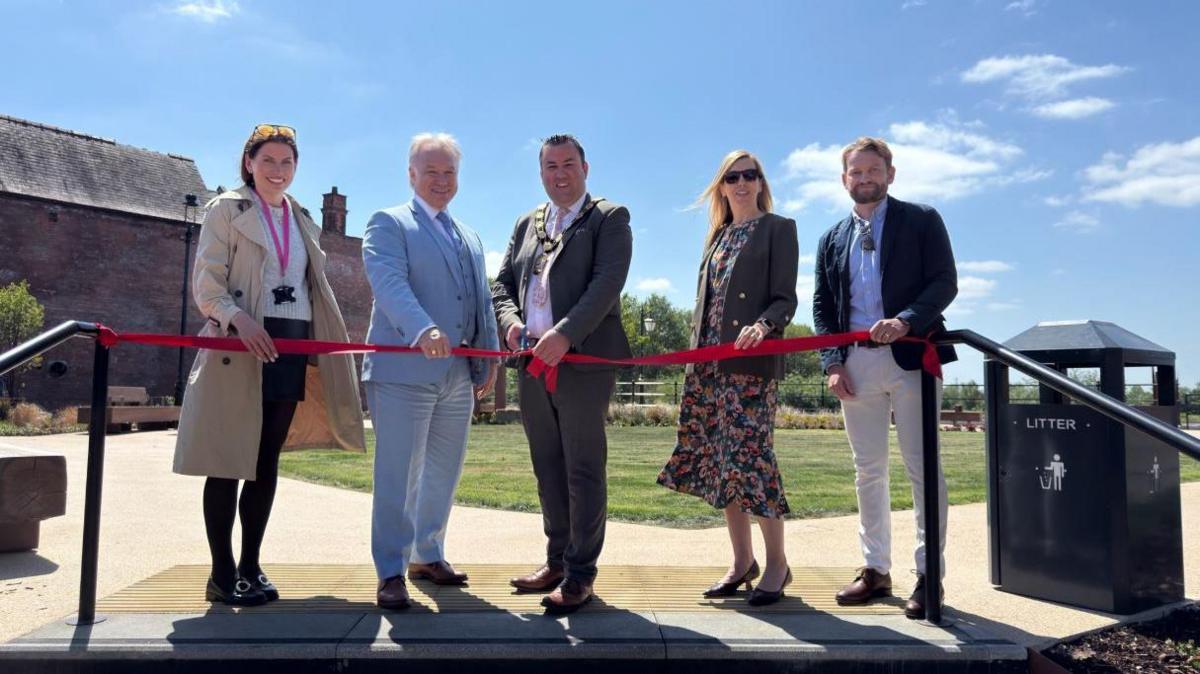 Five people standing outdoors, smiling and holding a red ribbon for a ceremonial cutting. The weather is sunny with a bright blue sky. Behind them is a landscaped area with grass and paths, and an old brick building on the left.