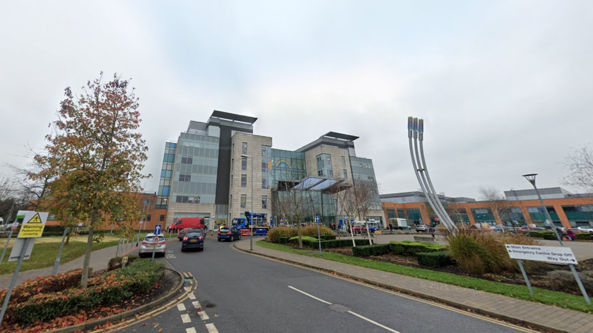 The outside of a hospital building with a glass front. There are cars parked outside the front entrance. 