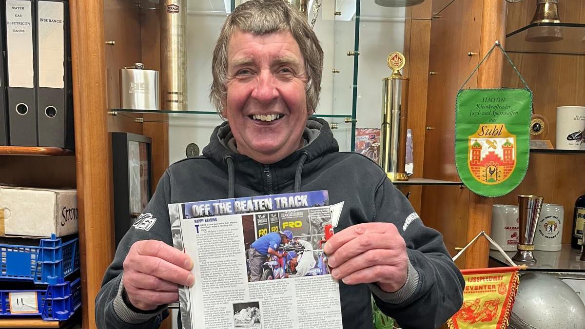 Ted Midgley stood up holding a magazine, which shows his article. He has a mod haircut and is wearing a black hoody.  