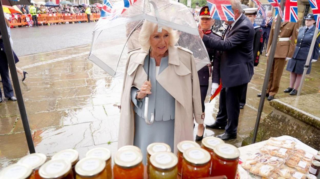 The Queen in a grey suit holding a clear plastic umbrella with a rain coat draped over her shoulders. There are crowds behind barriers behind her. There are jars and baked goods on a table in front of her. A man and woman in military uniform are standing behind her.