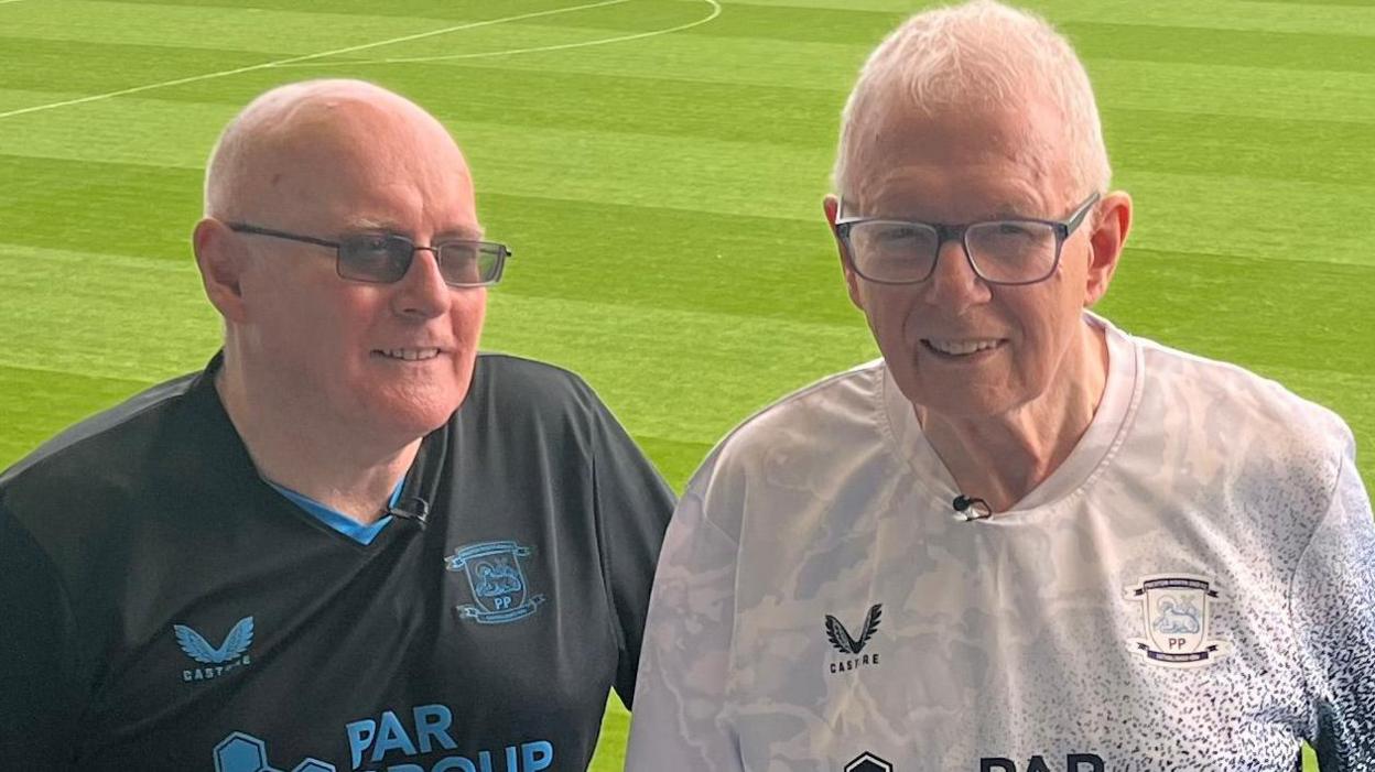 Peter and Terry Kilgariff are standing side by side in the stand at Deepdale Stadium - the home of Preston North End. They are both wearing Preston North End tops and the pitch can be seen behind them.