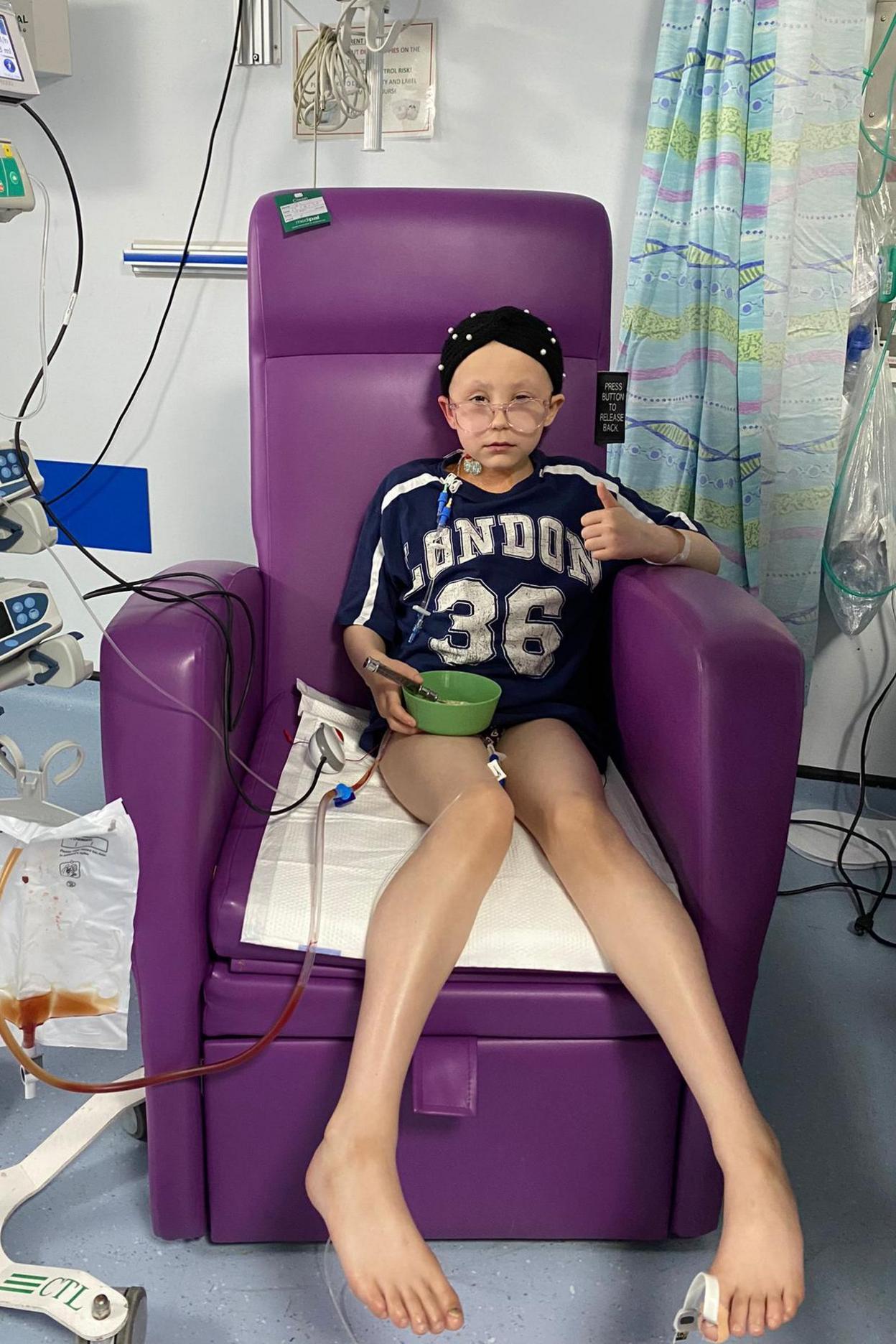 A  10 year old girl sits in a purple hospital armchair. She is wearing pyjama shorts and a t-shirt with a baseball style logo that reads London 36. She is wearing a black head scarf with white polka dots and glasses and is attached to medical machinery with tubes. She looks sad but is making a thumbs up with one hand and holding a green bowl in the other.