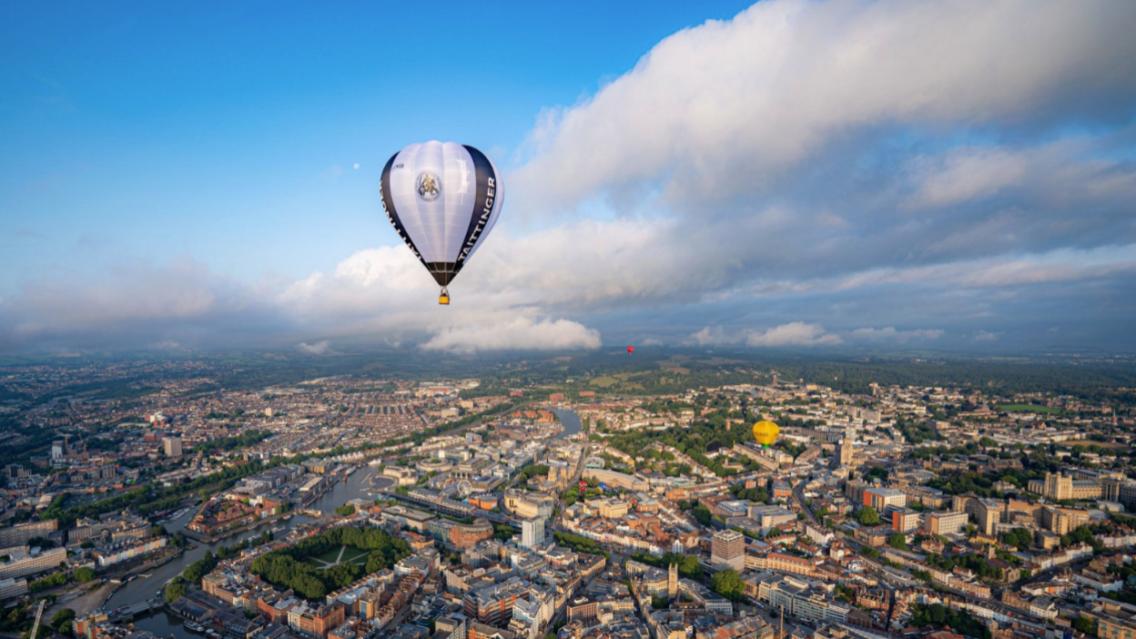A single hot air balloon is flying high above the city of Bristol. The river can be seen winding into the distance with rooftops and tall buildings visible all the way to the horizon. There are a few clouds in the sky and a few other balloons can be made out in the distance.