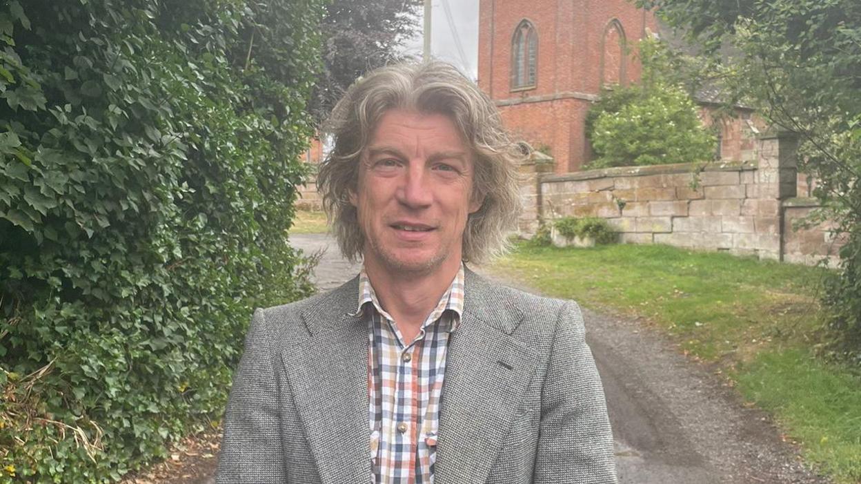 Scott wears a check shirt and jacket and is standing outside a church. 