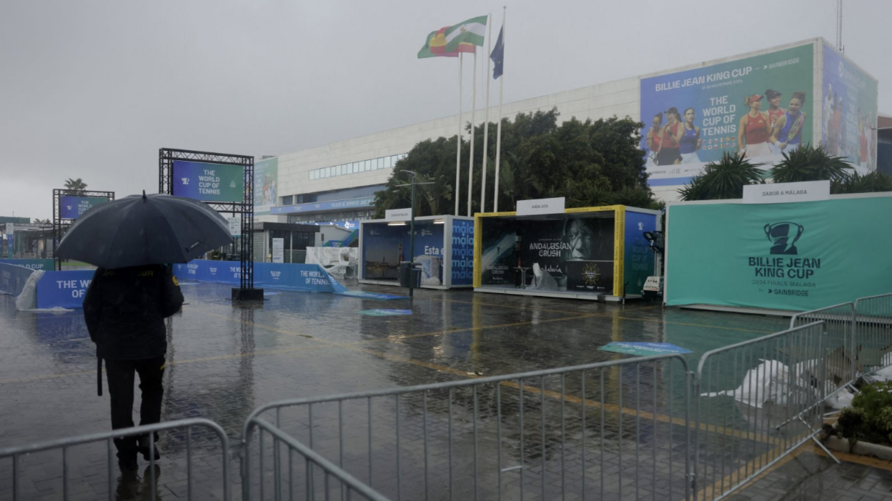 A man stands outside the Palacio de Deportes Jose Maria Martin Carpena Arena in Malaga holding a navy umbrella while torrential rain surrounds him. The man is looking at the tennis arena as a number of flags, including the Spanish and Andalusian flags, flutter in the wind