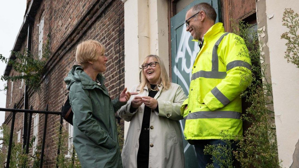 Tyne and Wear Building Preservation Trust chair Shona Alexander, Councillor Karen Kilgour, and Martin Hulse at the Keelmen's Hospital in Newcastle. They are standing in front of a green door covered in graffiti as they talk and smile. Ms Alexander is wearing a long green coat and has short blonde hair. Kilgour is wearing glasses, beige trench coat, black top and trousers. She has shoulder-length blonde hair. Mr Hulse is wearing glasses and a high visibility protective clothing. He has short grey hair.