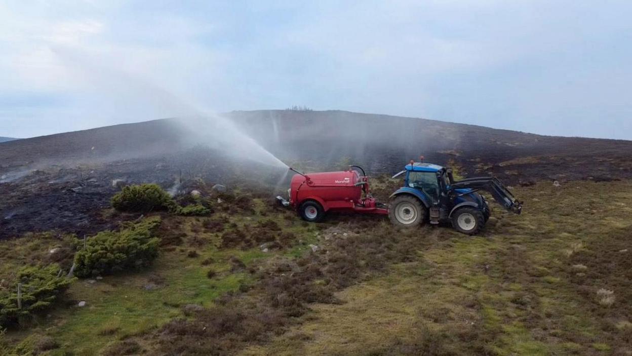 A red slurry tank attached to the back of a tractor sits on a moorland spraying water across an area of burnt land