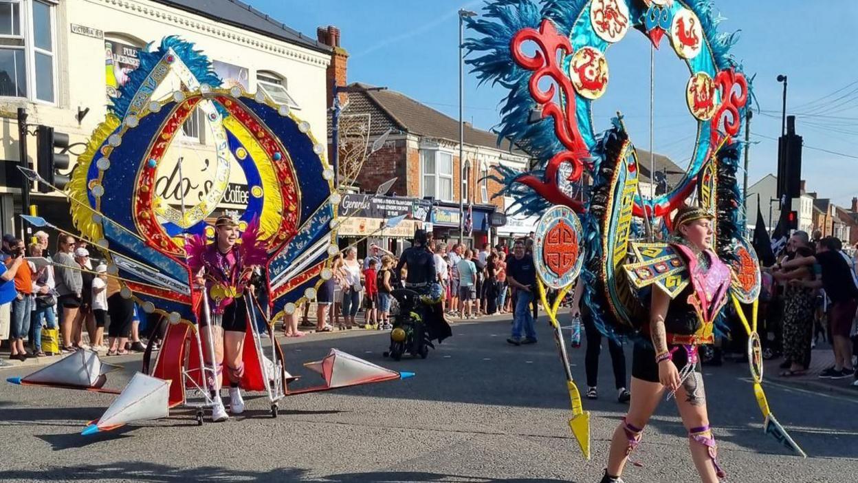 Two woman dressed in colourful carnival costumes walking along a street, There is a crowd of onlookers and there are shops along the road.