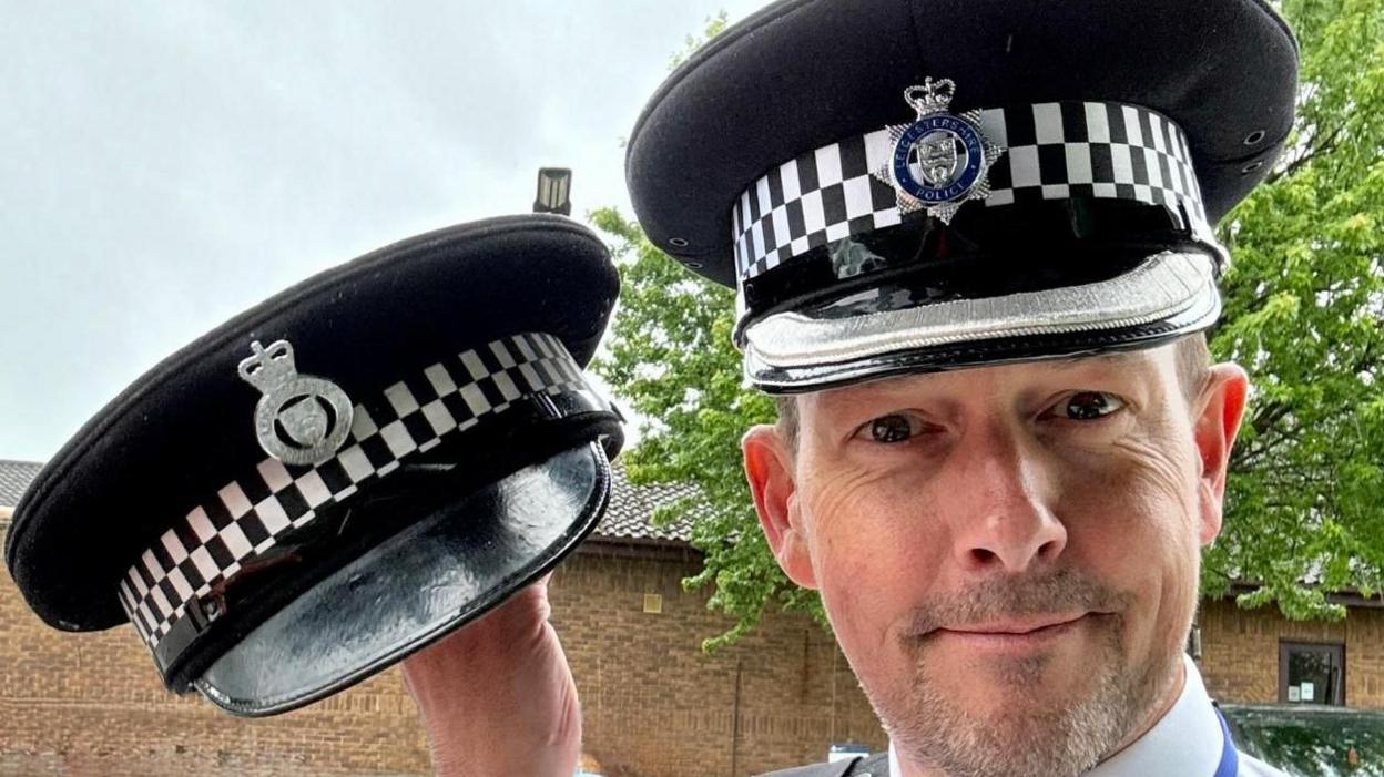 A policeman holding his lost hat up to the left of him. He is smiling and wearing his new hat also. There is a blue sky and a tree with green leaves in the background.