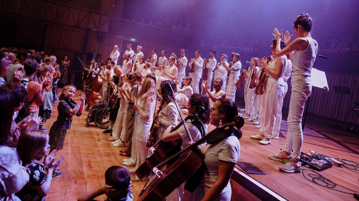 A collection of musicians on stage, all dressed in white, applauding