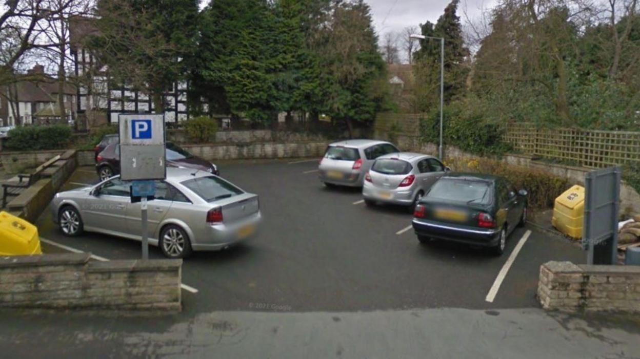 A small car park with nine spaces and five cars parked in it and a low brick wall in the foreground