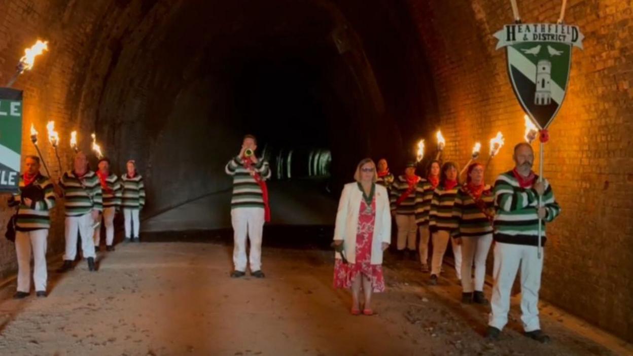 Lit torches being held in a disused railway tunnel in Heathfield in West Sussex. There is a trumpeter in the middle and people lined up along the edges of the tunnel holding torches and banners.
