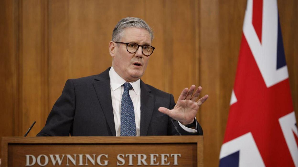 Sir Keir Starmer gestures from a Downing Street podium, wearing a navy suit and tie and a white shirt.