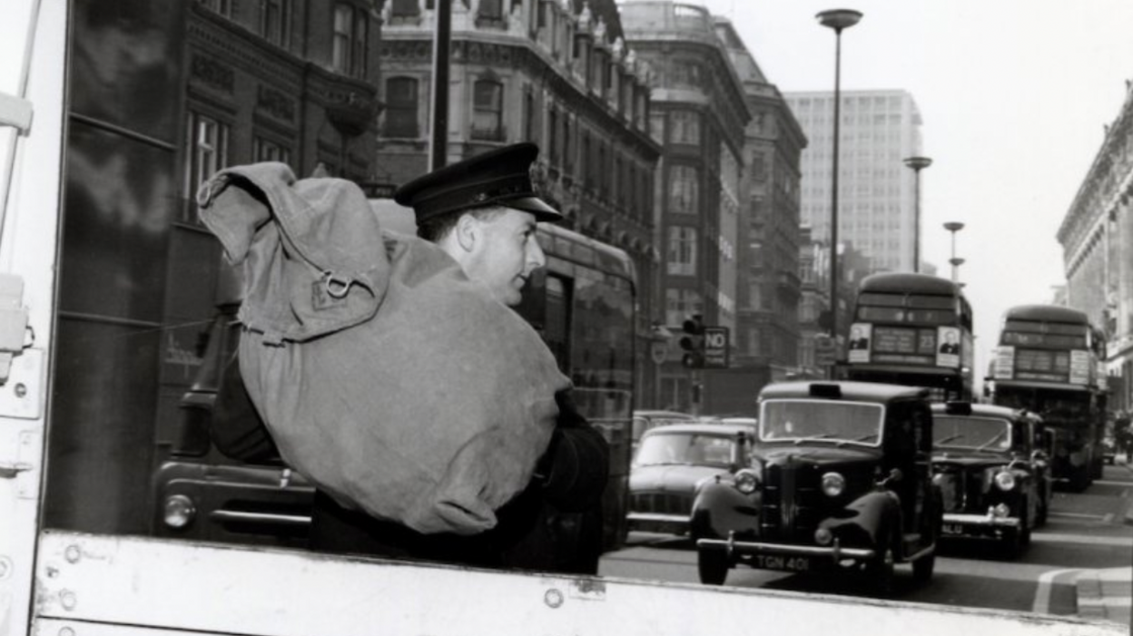 An old black and white photo of postman Rocky carrying his parcels in a sack on his back to hand-deliver them. Cars and iconic London red buses can be seen in the background