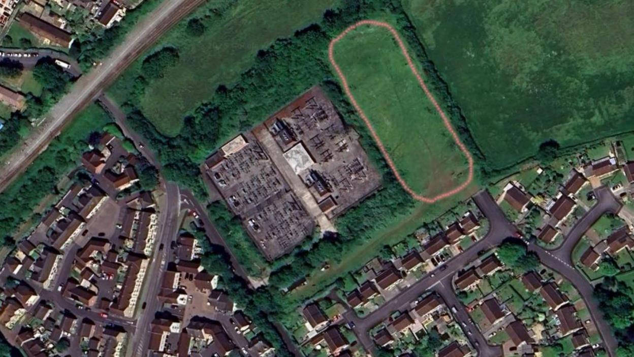 An aerial view showing where the proposed facility would have been built. There is a faint red line outlining a rectangular section of a field, and next to it is the existing electricity substation. There are lots of streets of residential housing nearby.