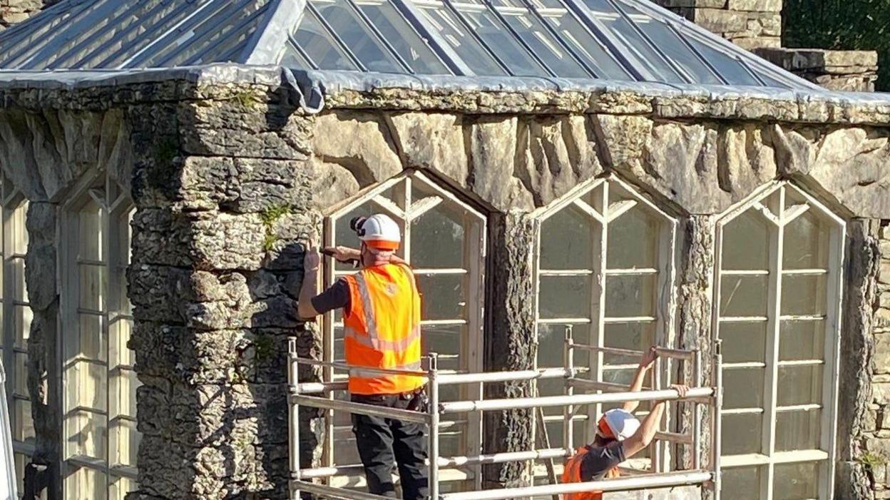 The Glasshouse which is an old, stone building with large pointed windows and a glass roof. Two workmen in orange hi-vis vests are on some scaffolding working on the windows.