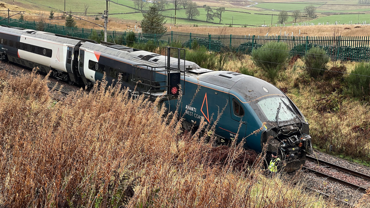 An Avanti West Coast train on a track in a rural setting. The front of the train is smashed and covered in mud.