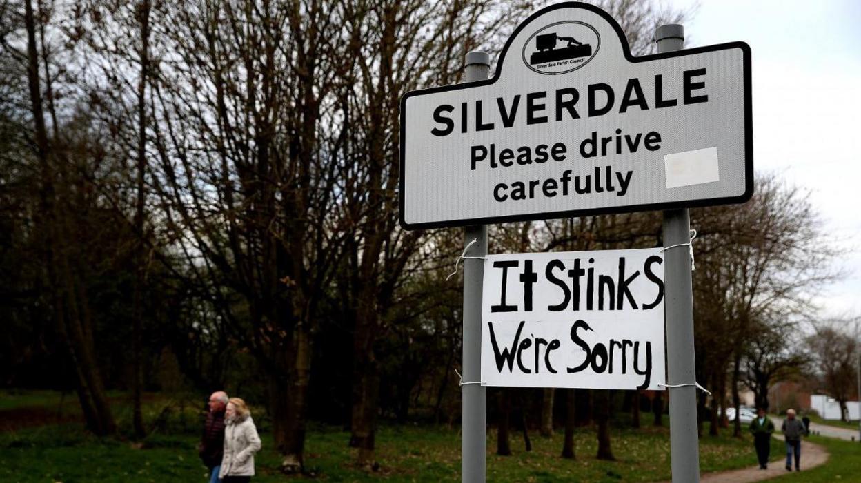 People walk past a sign in Silverdale asking motorists to drive carefully. Underneath, somebody has attached a poster reading: "It stinks, we're sorry." 