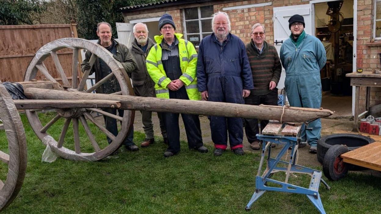 Five male volunteers stand next to the carriage, which is wooden and has two large wheels. One of the volunteers is in a high-vis yellow coat. 