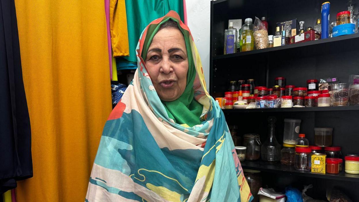 Howida Maher wearing a head scarf. It is long and goes around her shoulders. It is colourful. There are spices on a shelf behind her.