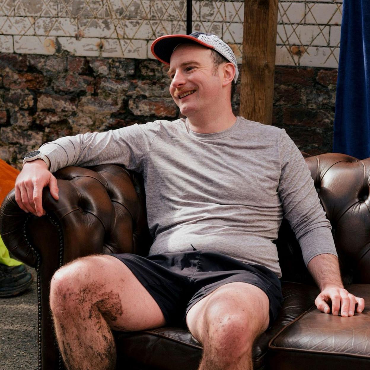 Xander, a 37 year old man is sat on a on a sofa in shorts and a running top. He's laughing and he's got a muddy leg