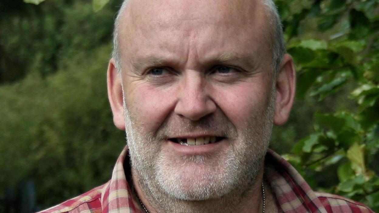 A head and shoulders picture of a man in a checked shirt with a light beard