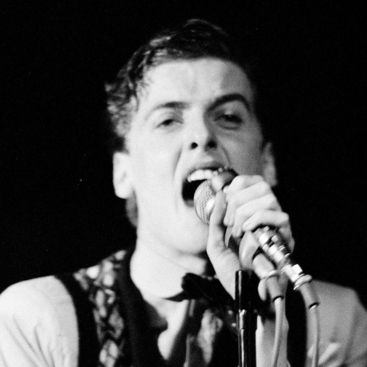 A black and white image of Peter Capaldi singing into a microphone while on stage in the late 70s or early 80s