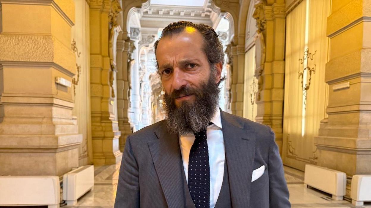 The lawyer, bearded, wearing a three-piece grey suit, white shirt and black white-dotted tie