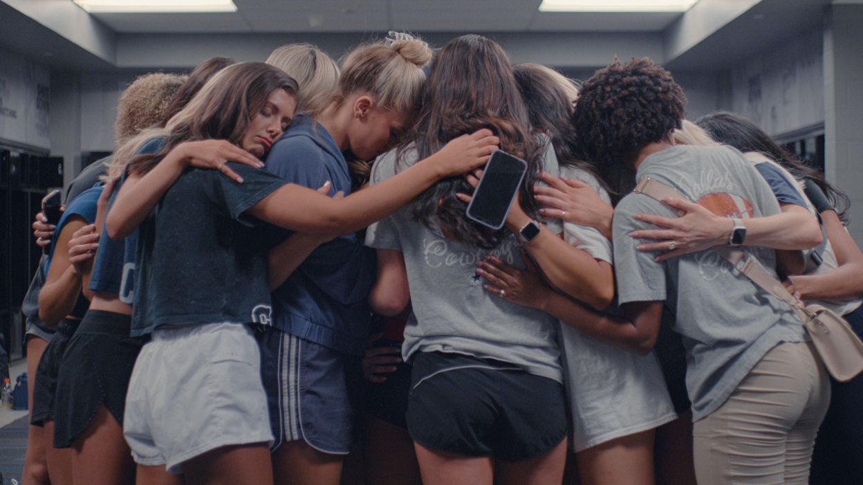 A large group of cheerleaders looking upset and consoling each other in a huddle