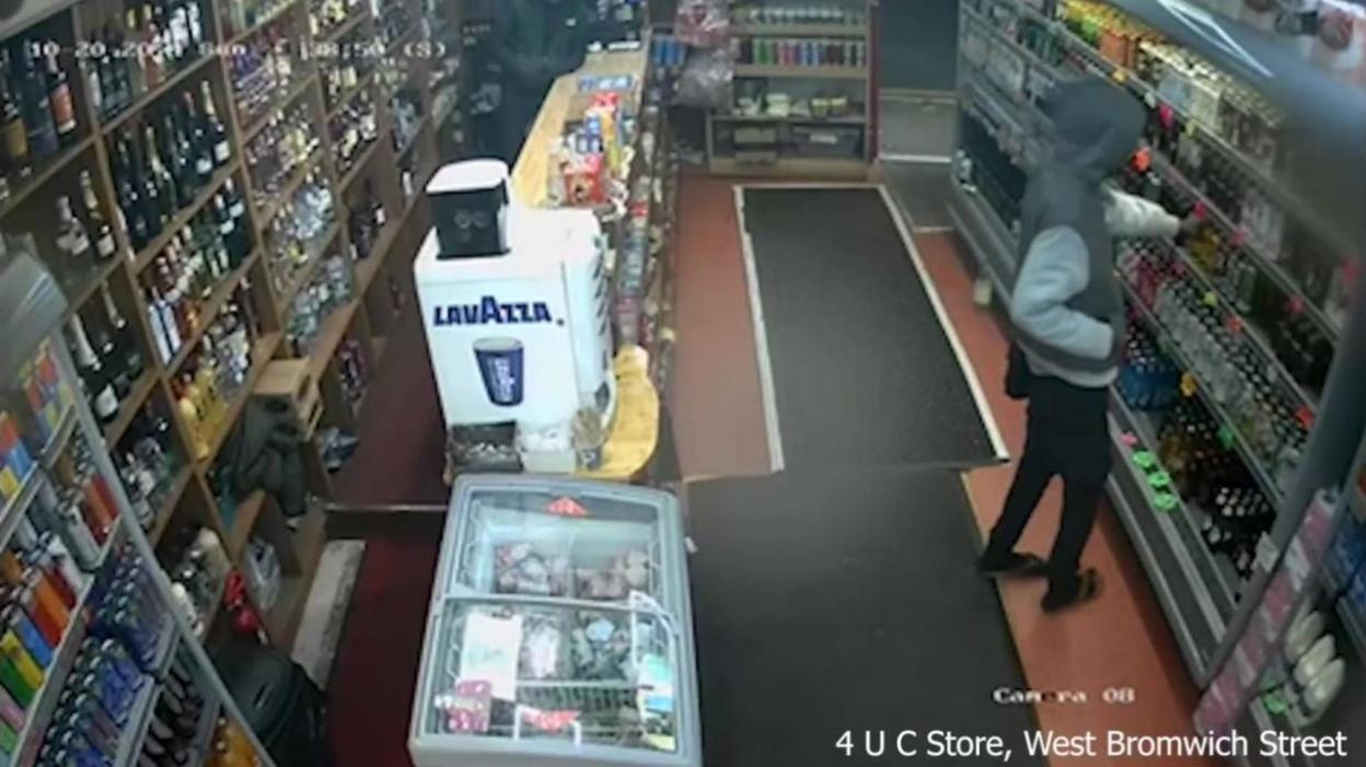 A CCTV image of a man in a grey top in a shop with rows of goods on both sides