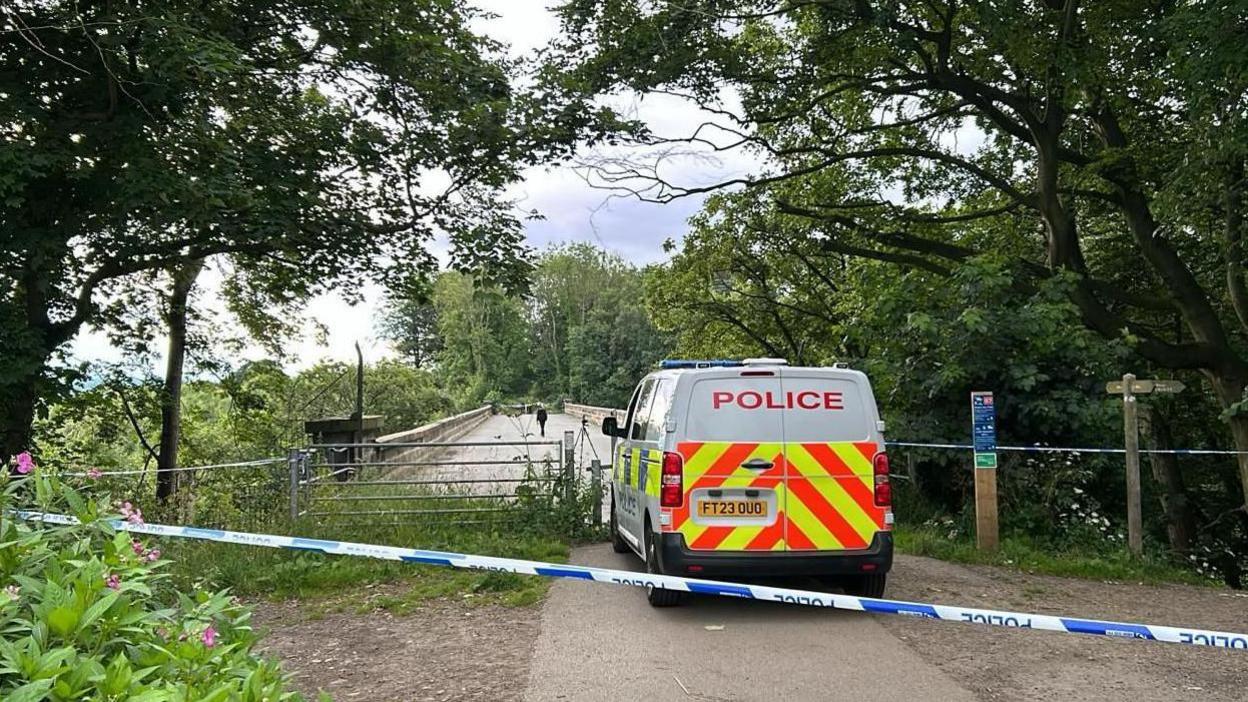 Police van in front of bridge with police tape behind it