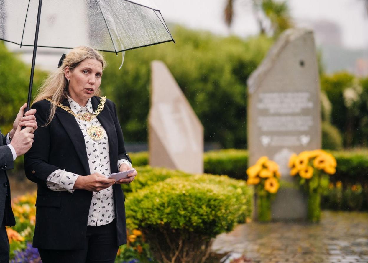 Natalie Byron Teare wearing a black jacket and the mayoral chain speaks to the crowd in front of the three-pillar stone Summerland memorial in a kept garden.