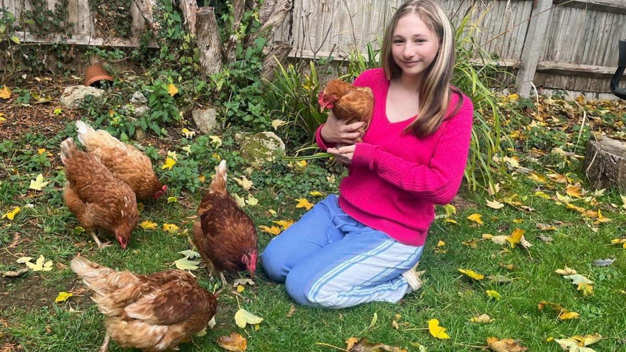 Poppy is wearing a bright pink jumper and light blue trousers. She is holding a brown hen and is kneeling on the grass. Four other brown hens are in front of her.