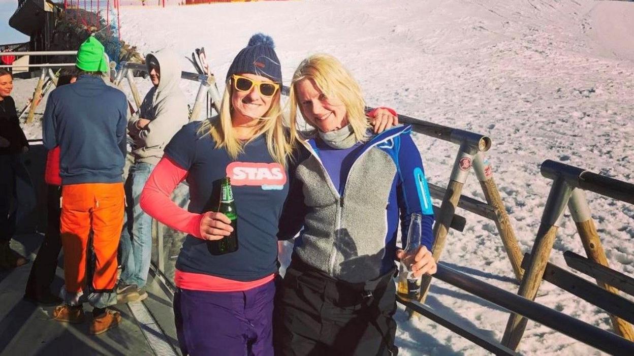 Susannah is wearing a wooly hat with a pom pom and holding a beer. She has her arm around a friend who has blonde hair and they are standing on the edge of a ski slope.