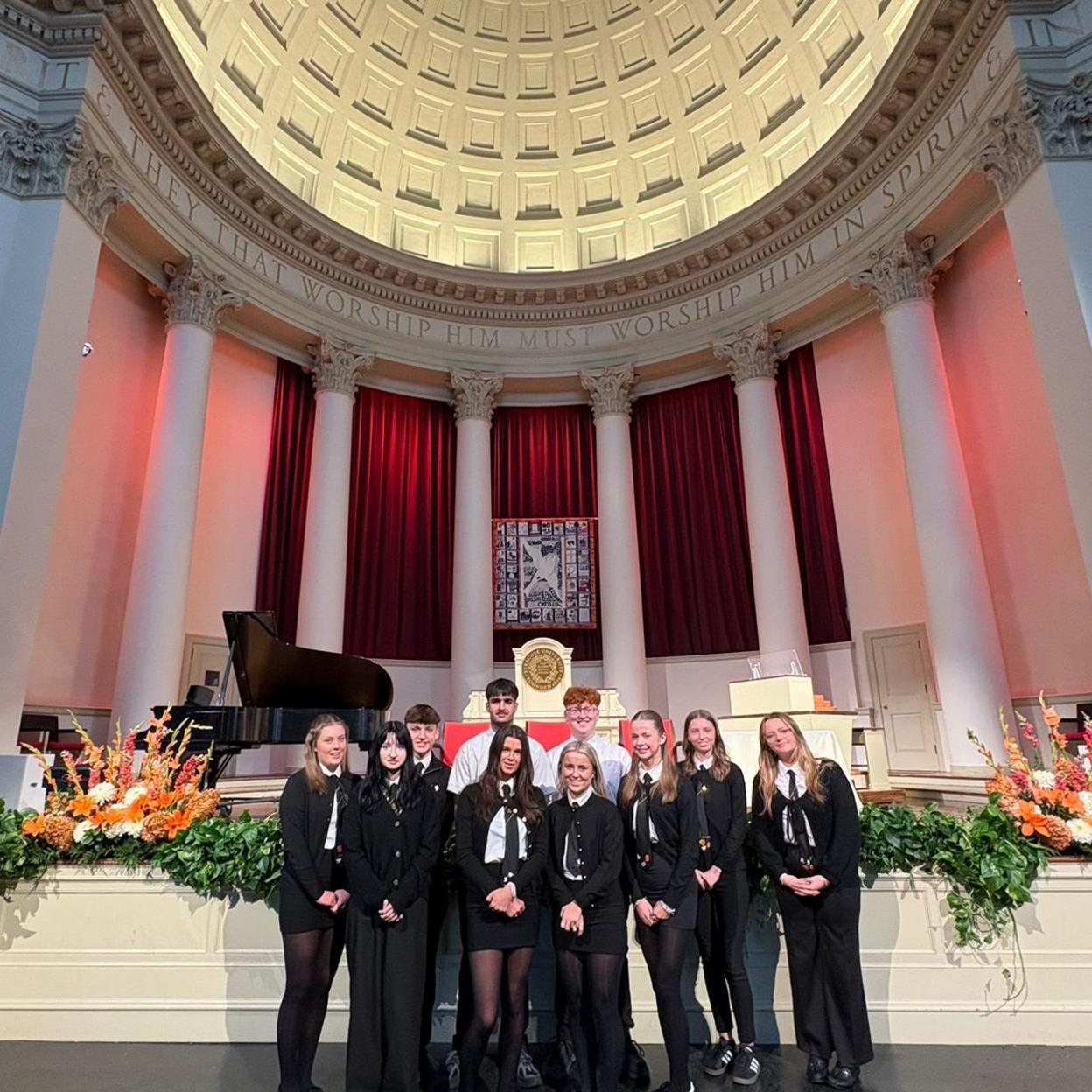Seven girls and three boys in school uniform, standing beneath a large dome in what appears to be a chapel. There are flower arrangements on either side of them and a grand piano in the background.