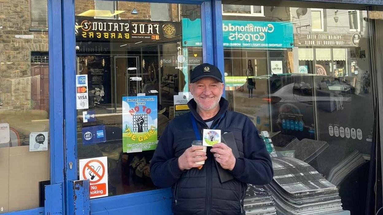 Photo of a man with a silver beard and a fred perry black baseball cap on. He stands outside a carpet store in the sunlight, holding a coffee and a "no to the gate" sticker. In the window behind him a "No to the gate" poster can be seen. 