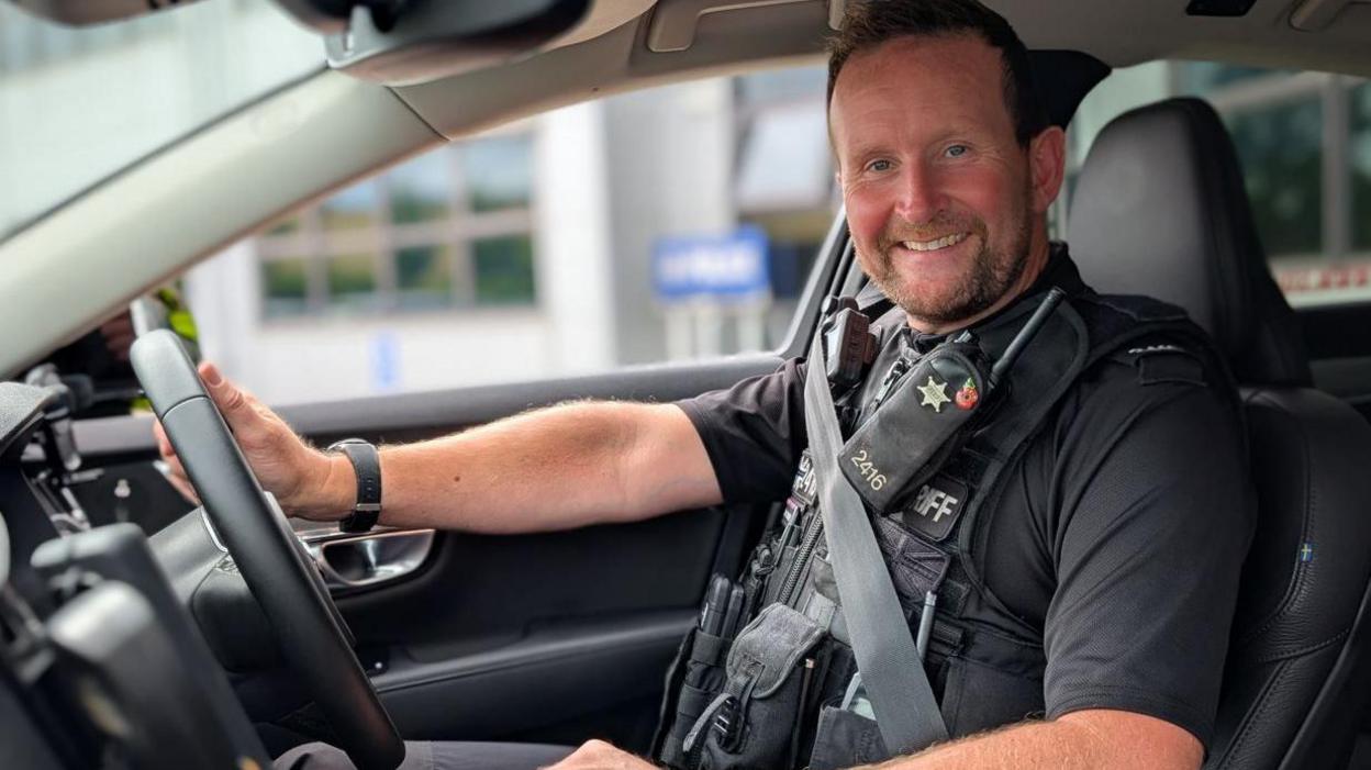 PC Connell is pictured sitting in a police car. He is smiling broadly and is wearing police uniform.