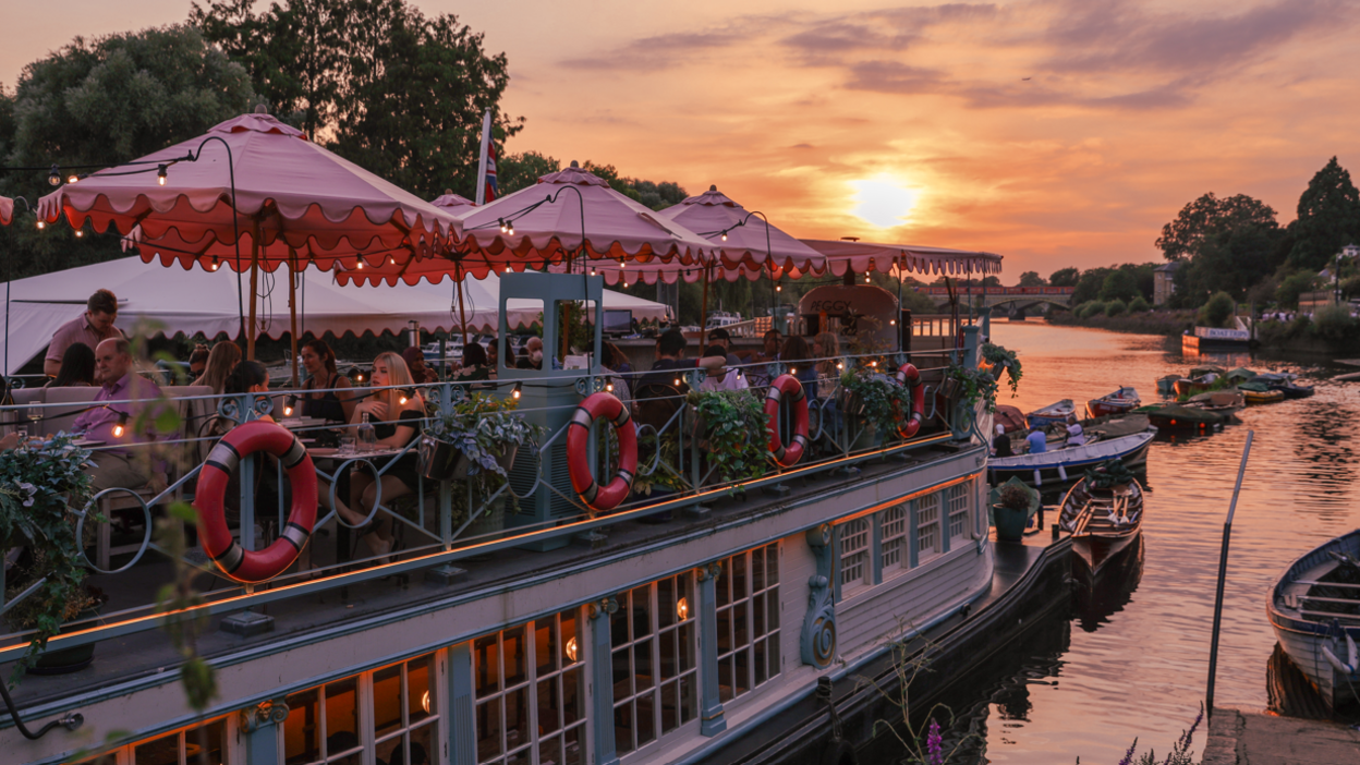 The Jesus College Barge, which is now Peggy Jean at Richmond-upon-Thames, full of diners on board at sunset. There are some smaller boats floating on the river nearby.