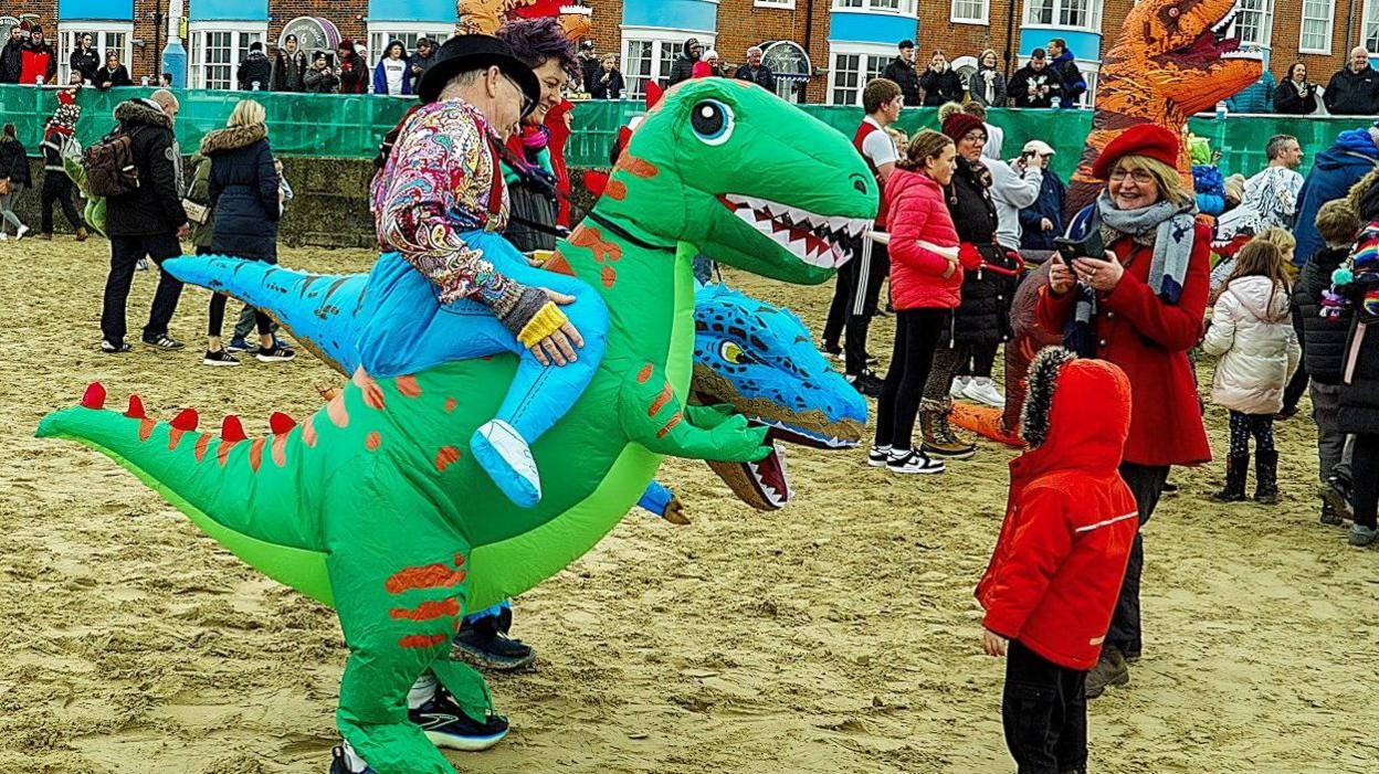 Two people dressed in dinosaur costumes talking to a young child wearing a red coat. They're standing on a beach and there's a crowd of people behind them.