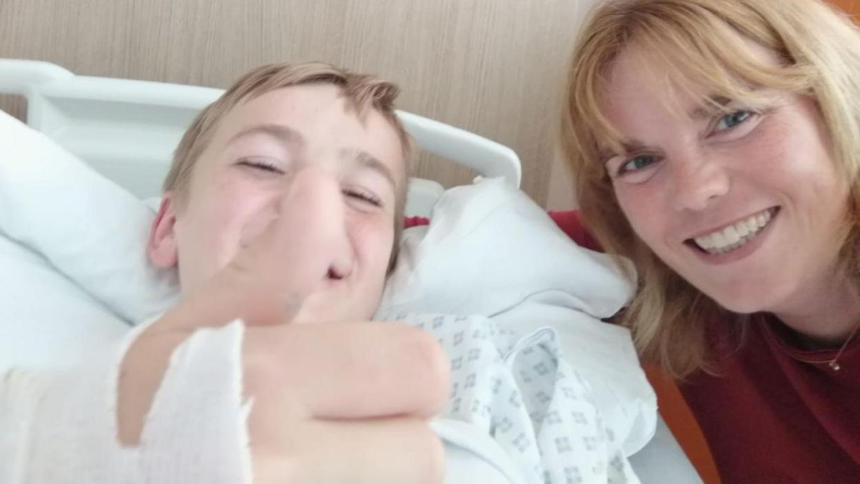 Alex has his thumb up in a hospital bed with his mum, Julie, next to him