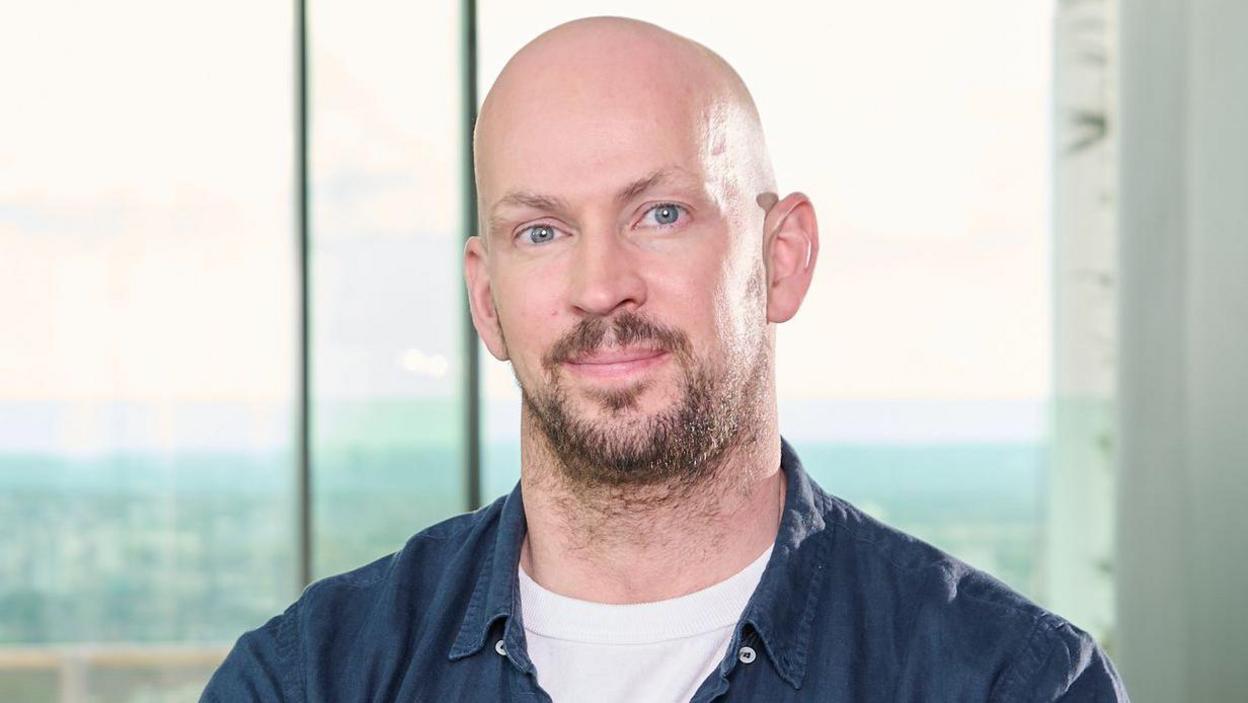 James Watt has a bald head and a beard. He is wearing a blue shirt over a white T-shirt and is sitting in front of a window
