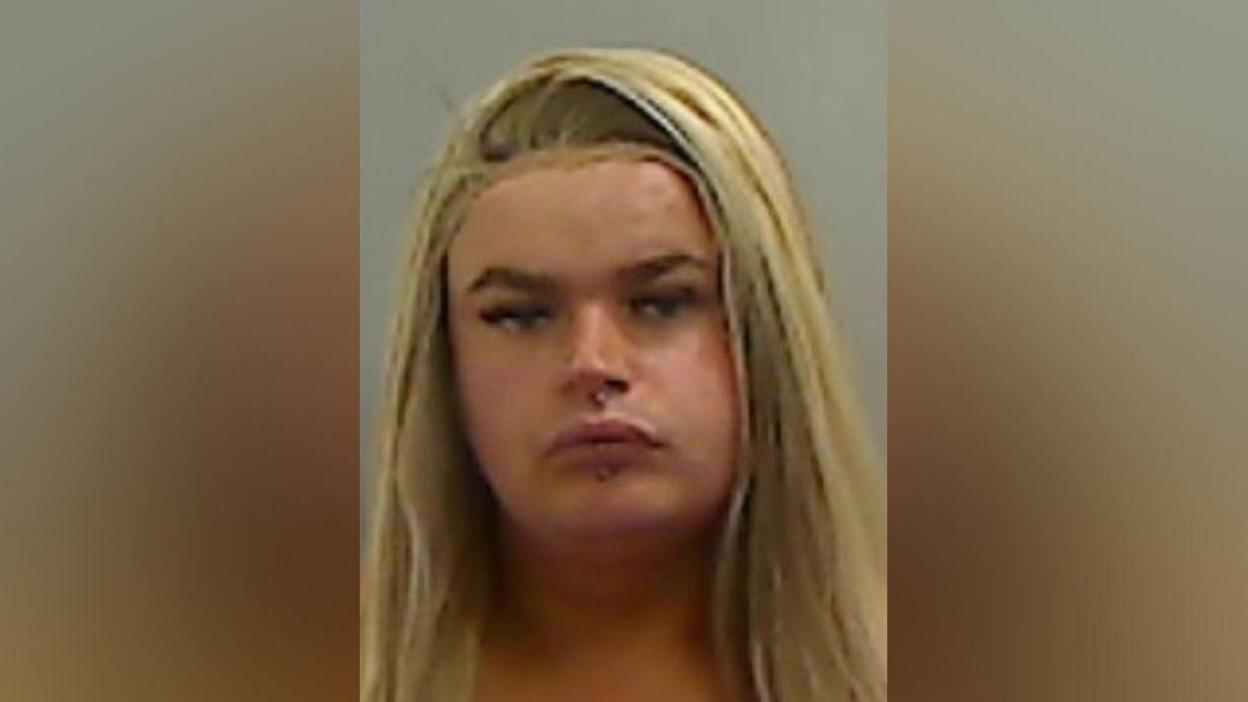 Mugshot of Watkin. She has long blonde hair, a full face of make up and piercings above and below her mouth.