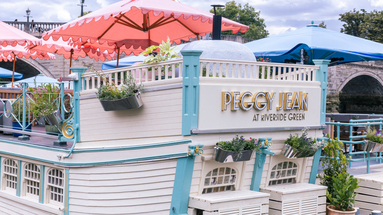View of the starboard of The Jesus College Barge, which is now restaurant Peggy Jean at Richmond-upon-Thames. The name of the restaurant is written on the vessel. There are several pots with green plants on board and red parasols.