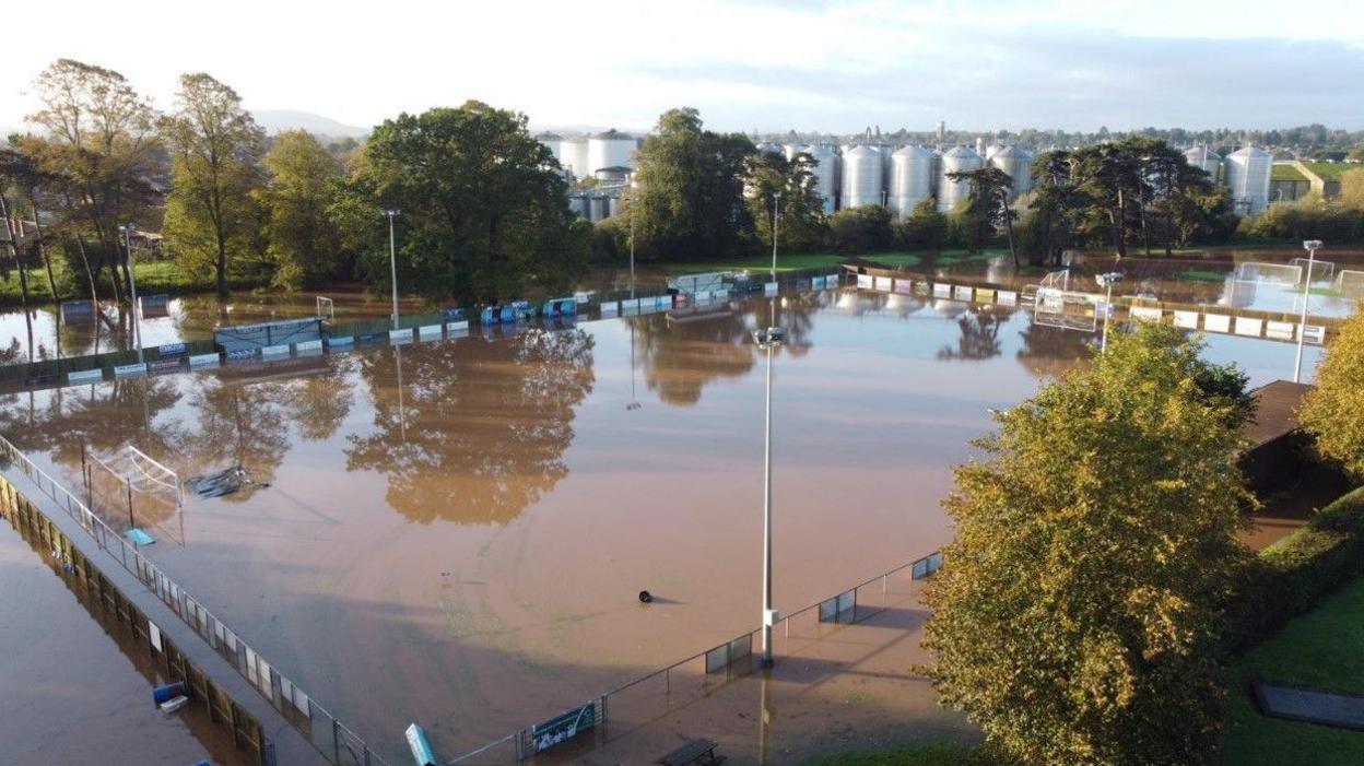 A football pitch covered in brown floodwater. The pitch is surrounded by trees
