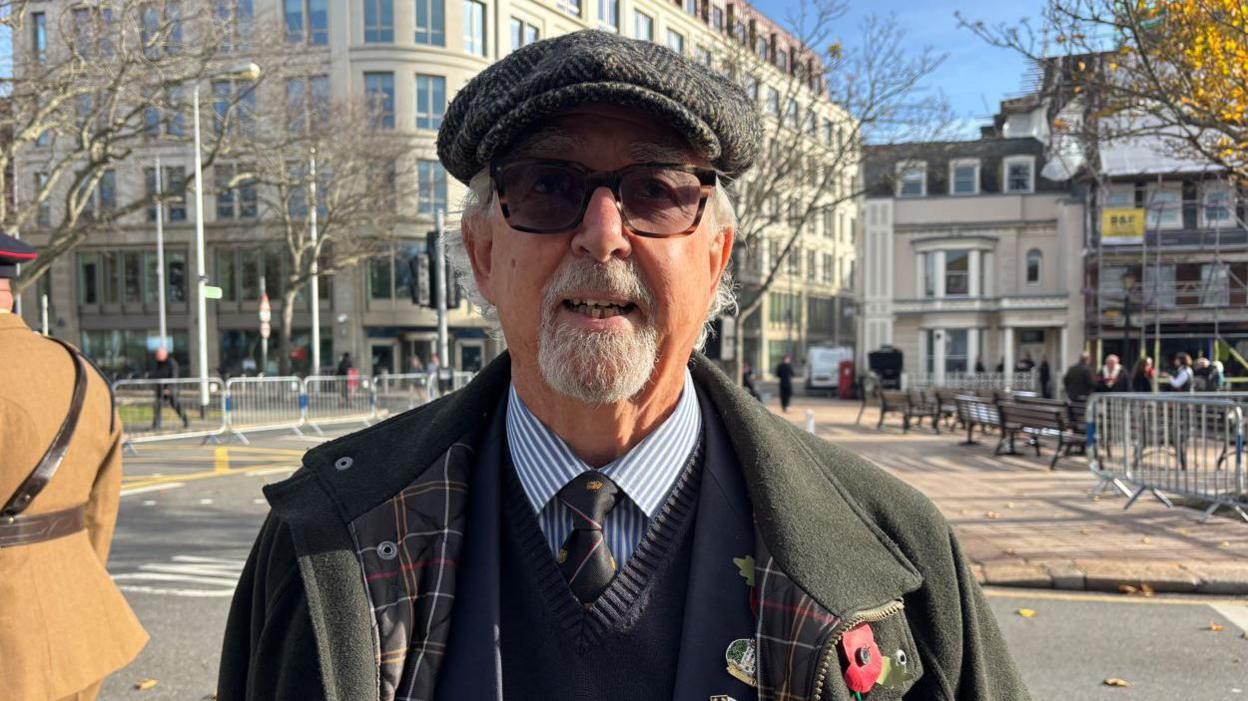 David Drage is in a green coat with a blue jacket shirt and tie underneath. He is wearing a hat and glasses and is smiling at the camera.