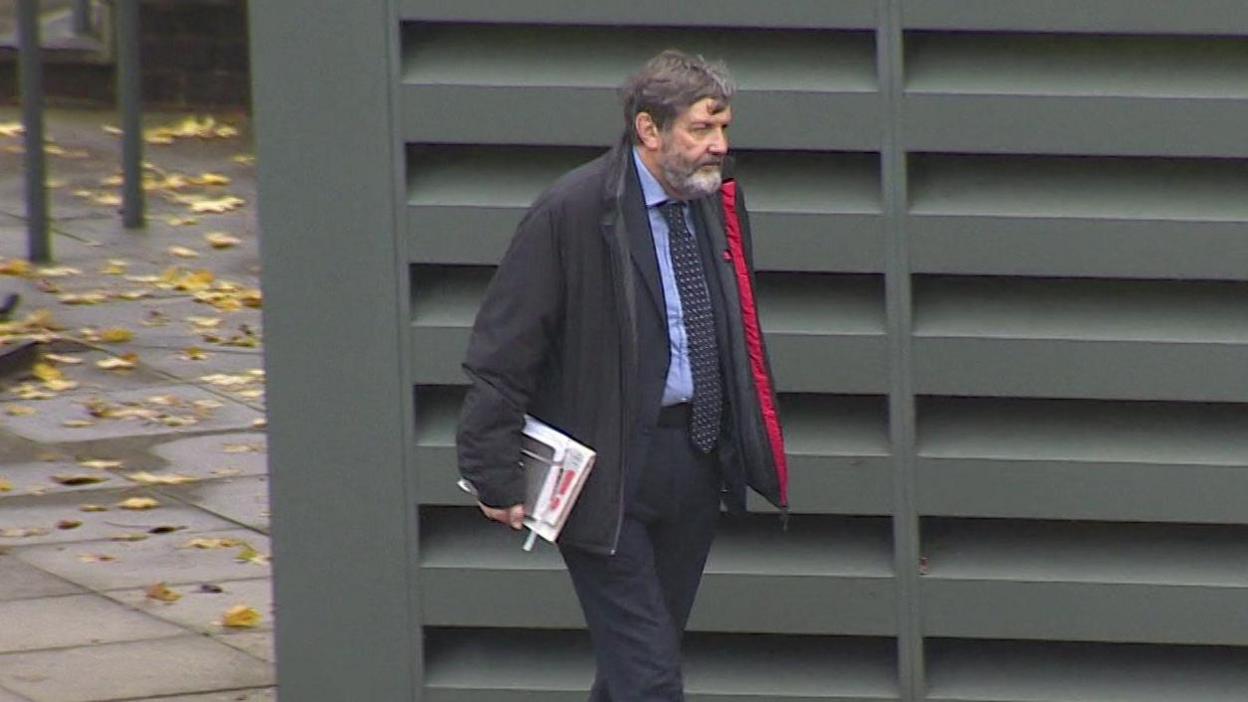 Alan Harris wearing a black coat with a red inner lining, dark suit, blue shirt and a dark blue tie with yellow markings on it walks past a green metal structure which has ventilation slots on the exterior. He is carrying a couple of notepads. Mr Harris has a greying beard and greying hair.
