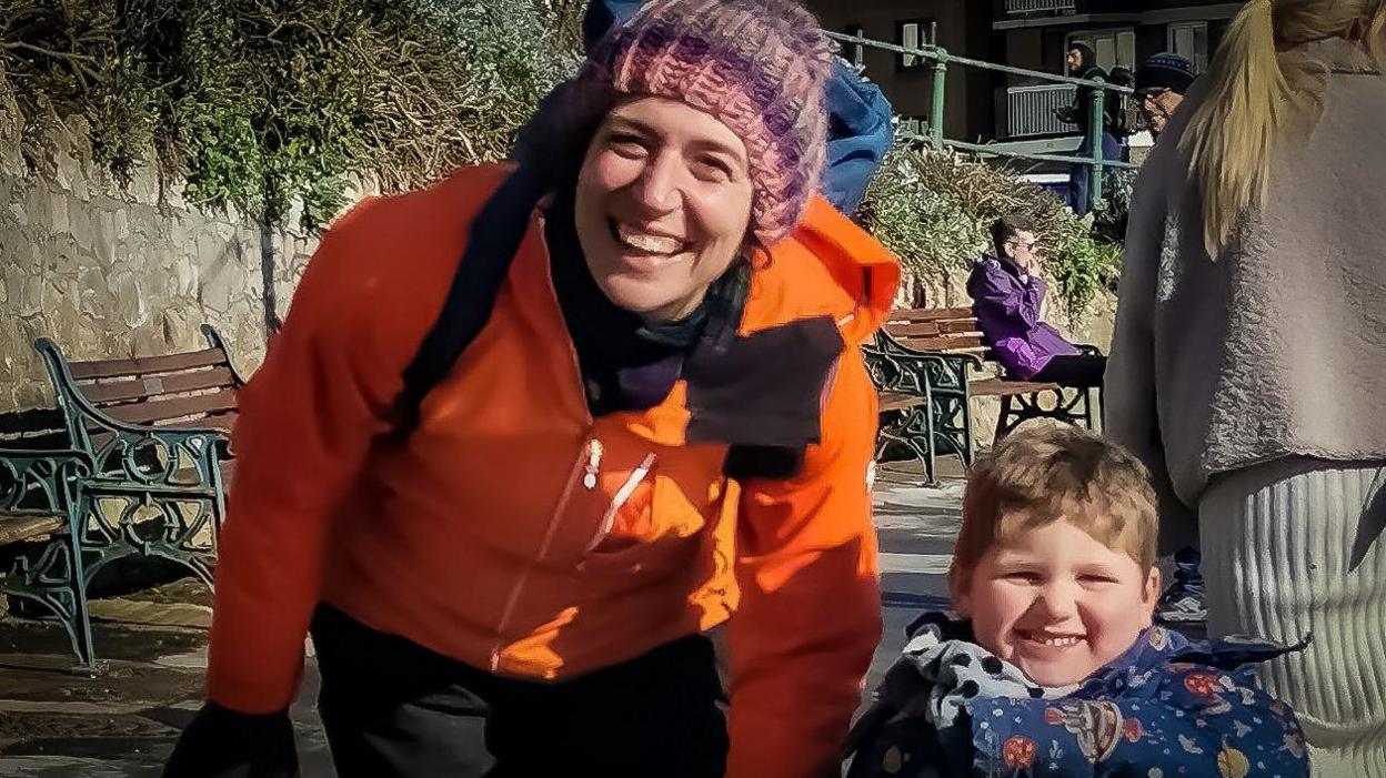 Sarah and Ivor are walking on a city street. Both are looking at the camera and smiling. They both have on winter coats