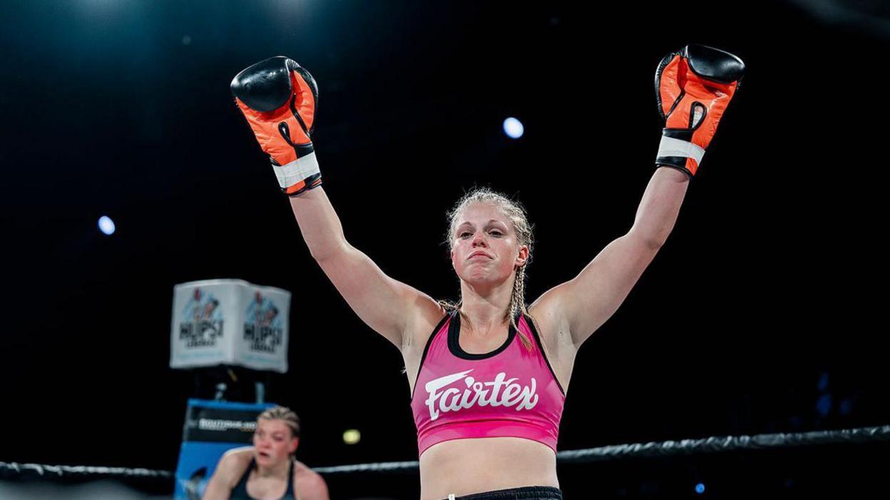 Hannah Turner, in a pink top, with her hands in orange and black boxing gloves raised after her win in Germany