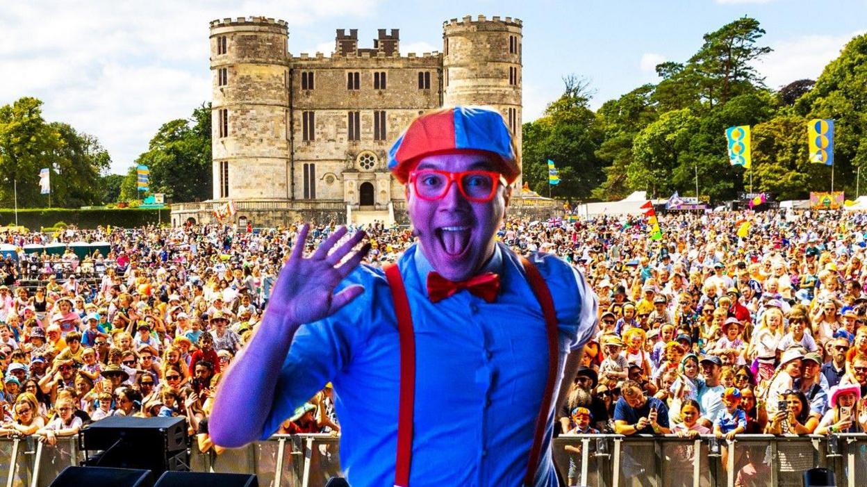 Blippi smiles at the camera with his mouth wide during his performance. Behind him is a huge crowd and in the distance is the stone crenelated keep of Lulworth Castle.